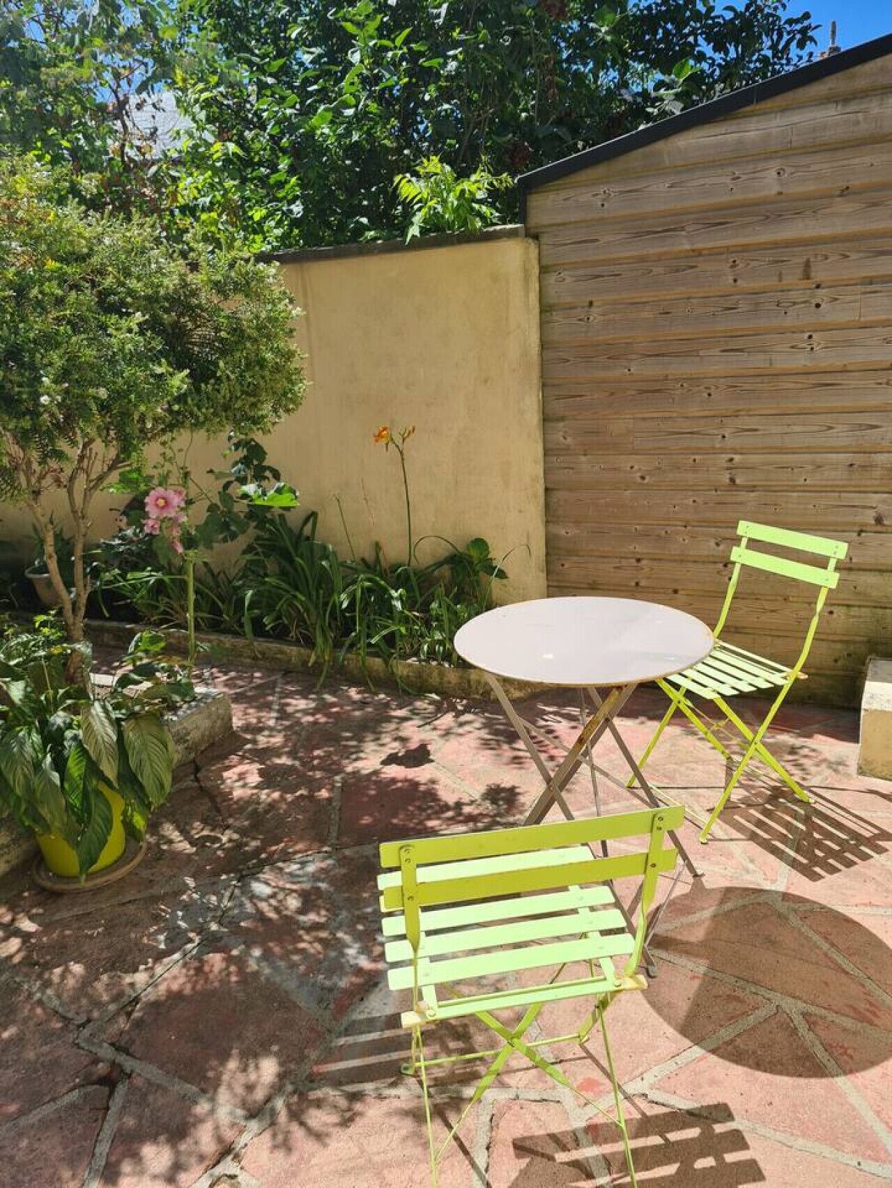 A patio with a round table and two green chairs, surrounded by potted plants and a wooden wall.