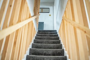 Un escalier avec des rampes en bois et des marches recouvertes de moquette, menant à une pièce dotée d’un radiateur et d’un tableau encadré accroché au mur.