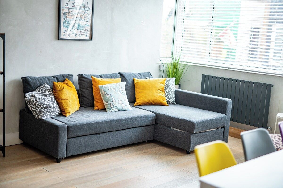Ein graues Ecksofa mit gelben und gemusterten Kissen, eine gerahmte Karte an der Wand und ein Fenster mit Jalousien.