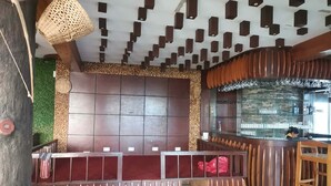 Un espace bar avec un comptoir en bois, un coin salon avec un banc rouge rembourré, et un mur orné d’un motif géométrique composé de panneaux rectangulaires sombres.
