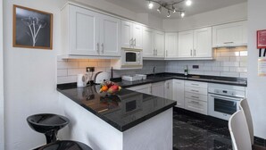 A modern kitchen with white cabinets, a black countertop, a microwave, and a fruit bowl. - Castle Harbour 39 - Two Bed (Los Cristianos)