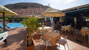 A poolside area with tables and chairs, a pool, and a view of a hillside town. - Castle Harbour 39 - Two Bed (Los Cristianos)
