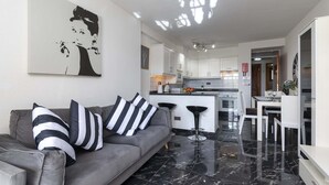 A modern living room with a grey sofa, black and white striped pillows, a black and white abstract painting on the wall, and a kitchen in the background. - Castle Harbour 39 - Two Bed (Los Cristianos)