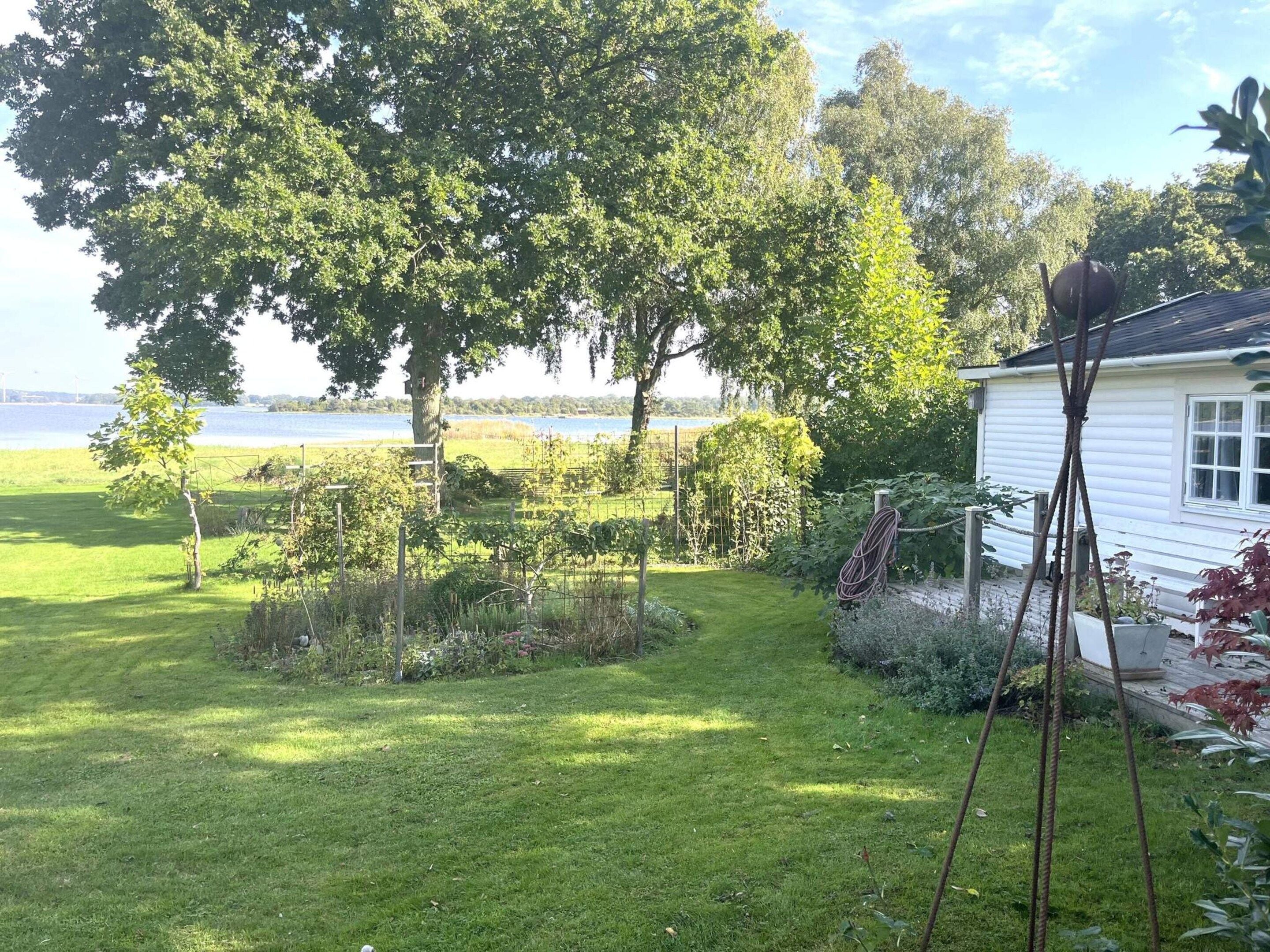 A white house with a garden in front, a body of water in the background, and a clear sky.