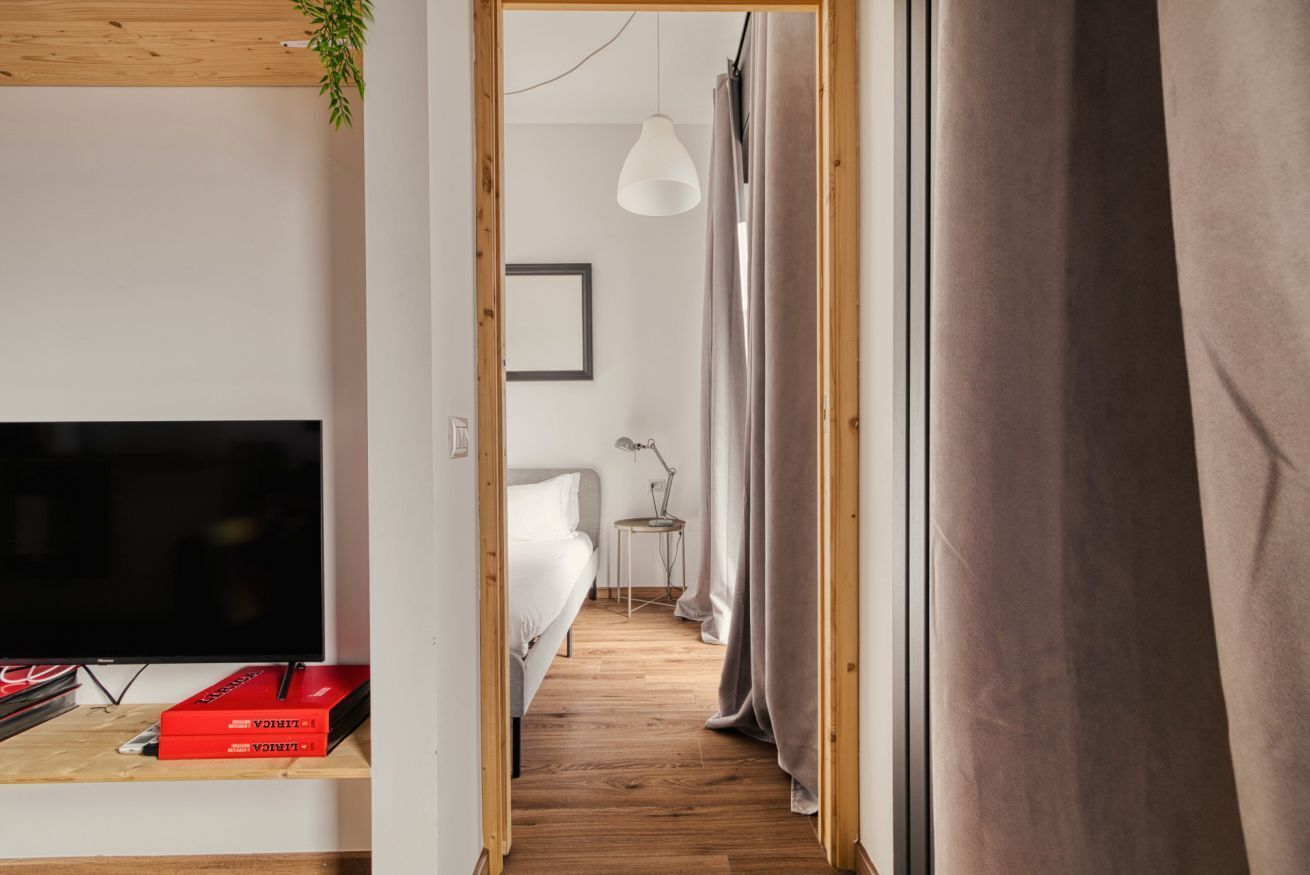 A narrow hallway with a wooden floor, a bed with white bedding, a bedside table with a lamp, and a television on a stand.