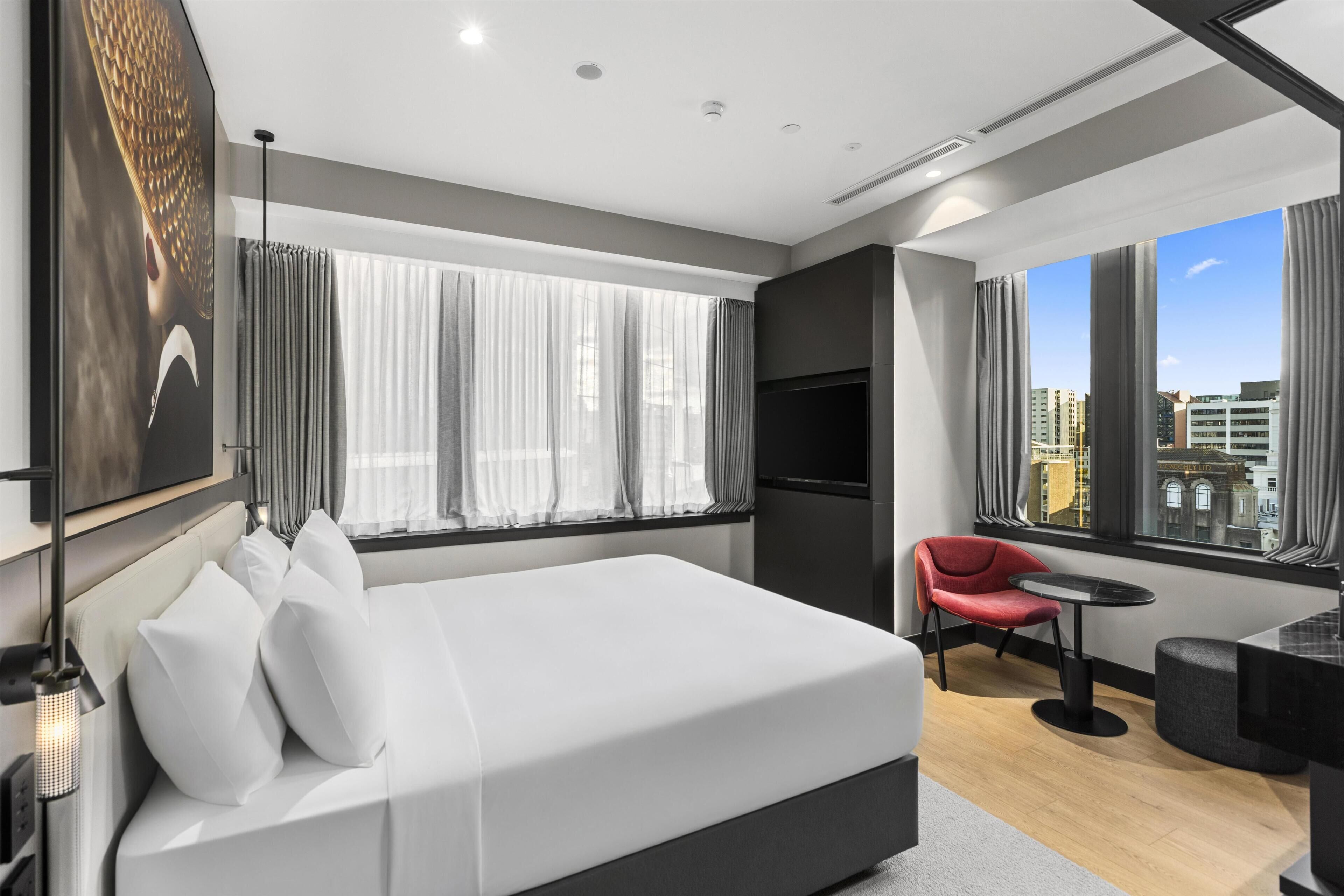 A modern hotel room with a large bed, a red chair, a small table, and a city view through large windows.