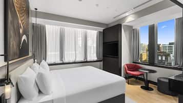 A modern hotel room with a large bed, a red chair, a small table, and a city view through large windows.