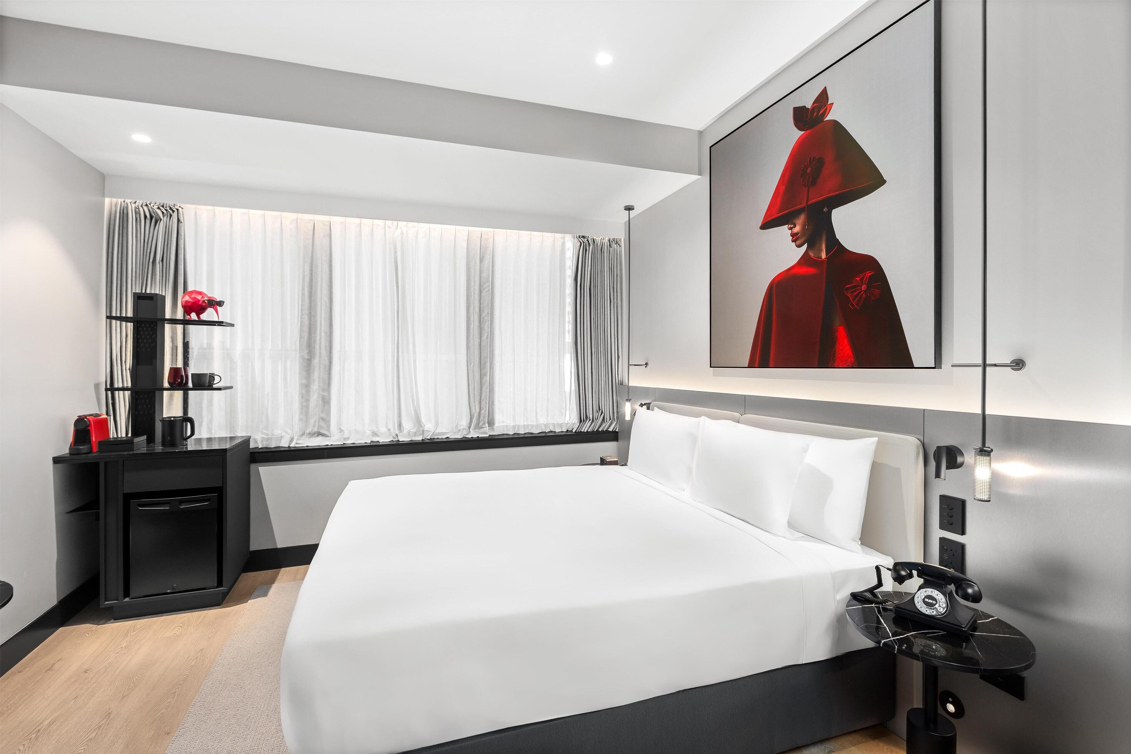 A modern hotel room with a large bed, a bedside table with a telephone, a small desk with a computer and a red object, and a window with curtains.