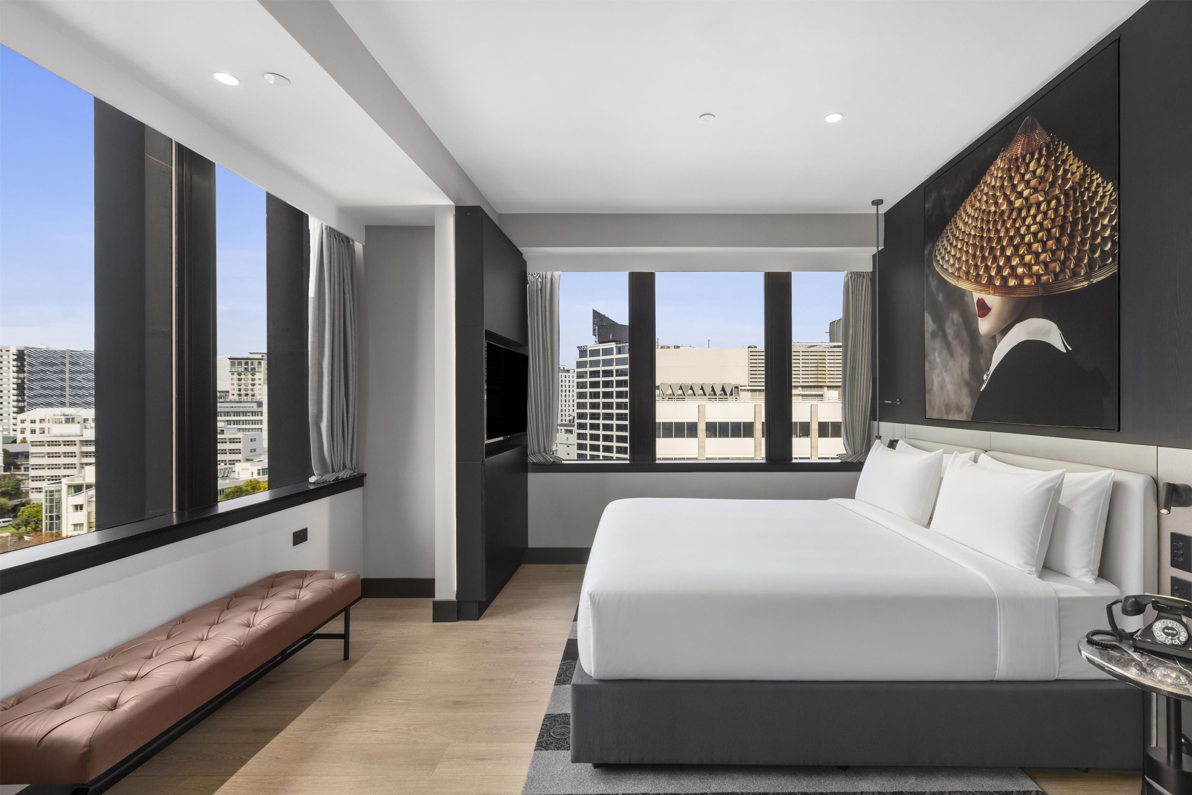 A modern hotel room with a large bed, a bench, a flat-screen TV, and a city view.