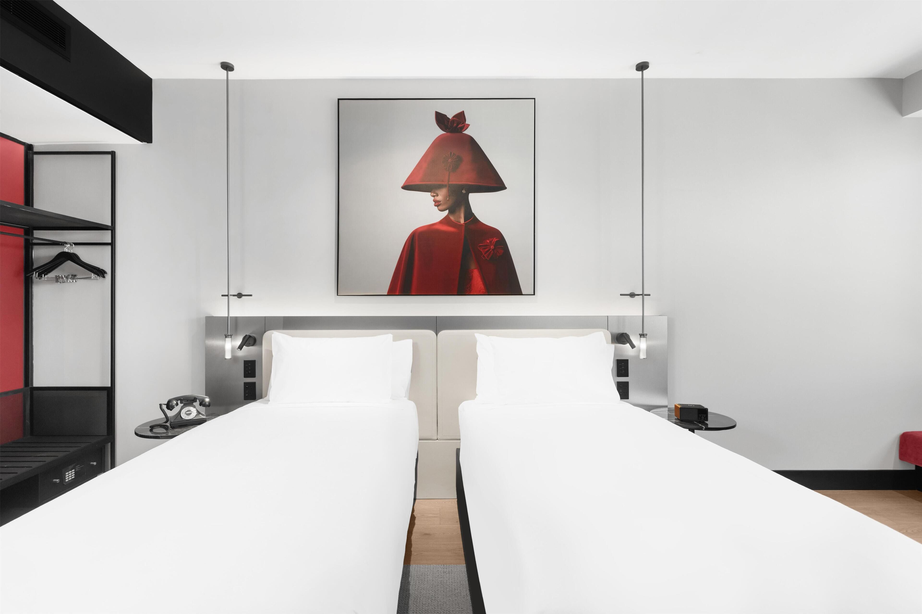 A modern hotel room with two single beds, a large framed artwork on the wall, a red coat hanging on the wall, and a small table with a telephone and a lamp.