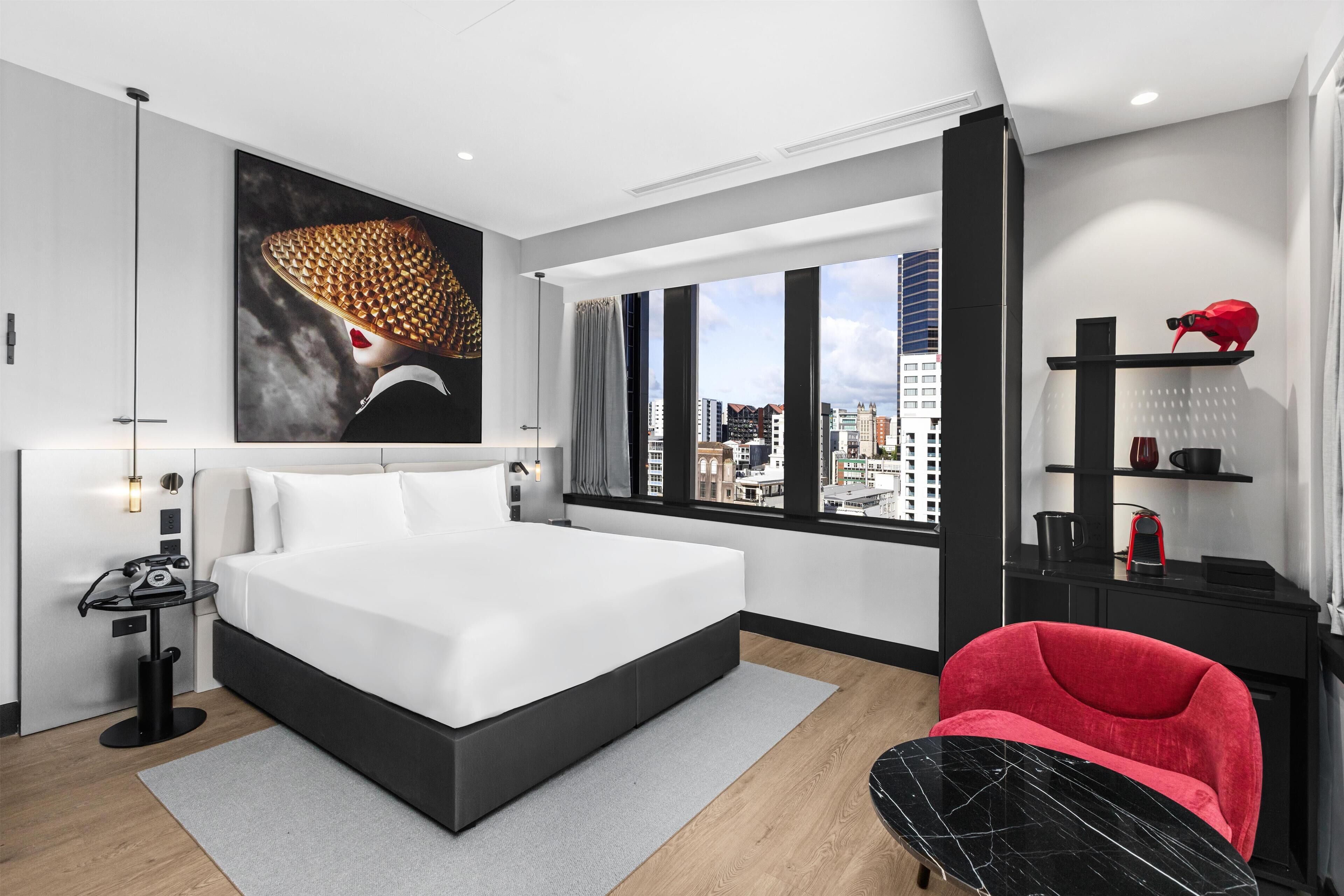A modern hotel room with a large bed, a city view, a red chair, and a black coffee table.