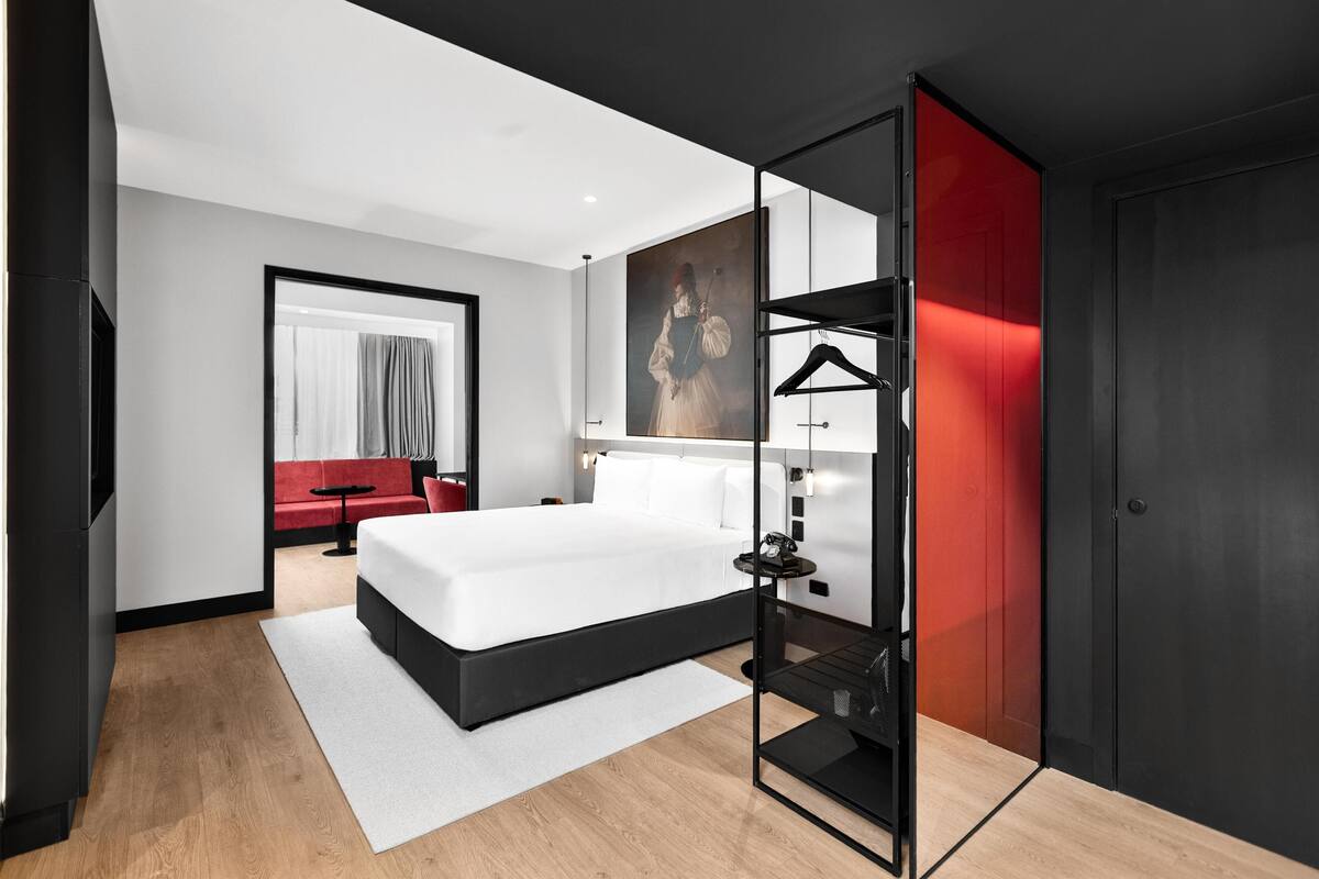 A modern hotel room with a large bed, a red sofa, a painting on the wall, and a red door.