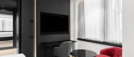 Habitación de hotel moderna con un televisor de pantalla plana grande, una mesita redonda pequeña, dos sillas y un ventanal con cortinas translúcidas.