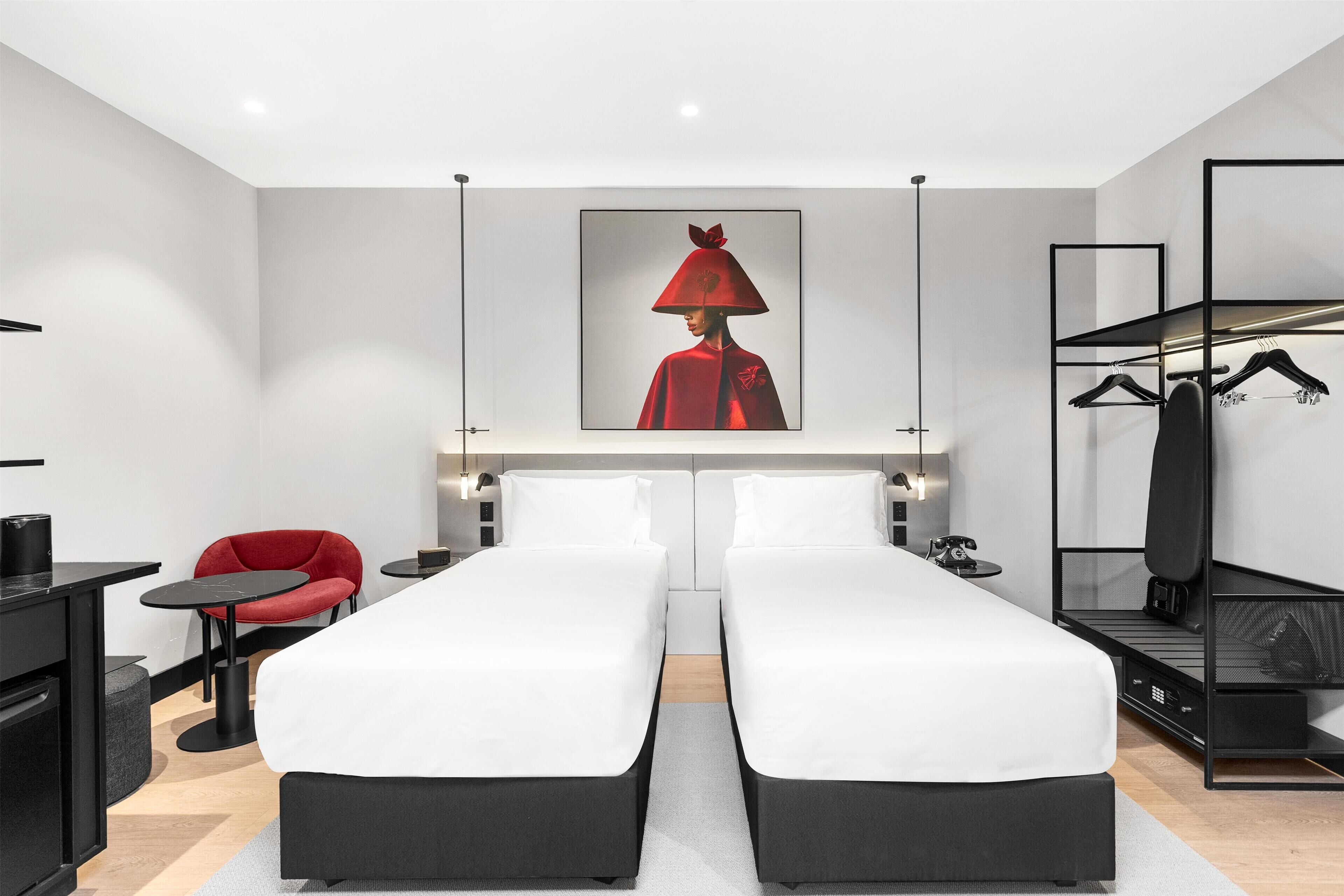 A modern hotel room with two beds, a red chair, a black desk, and a coat rack.