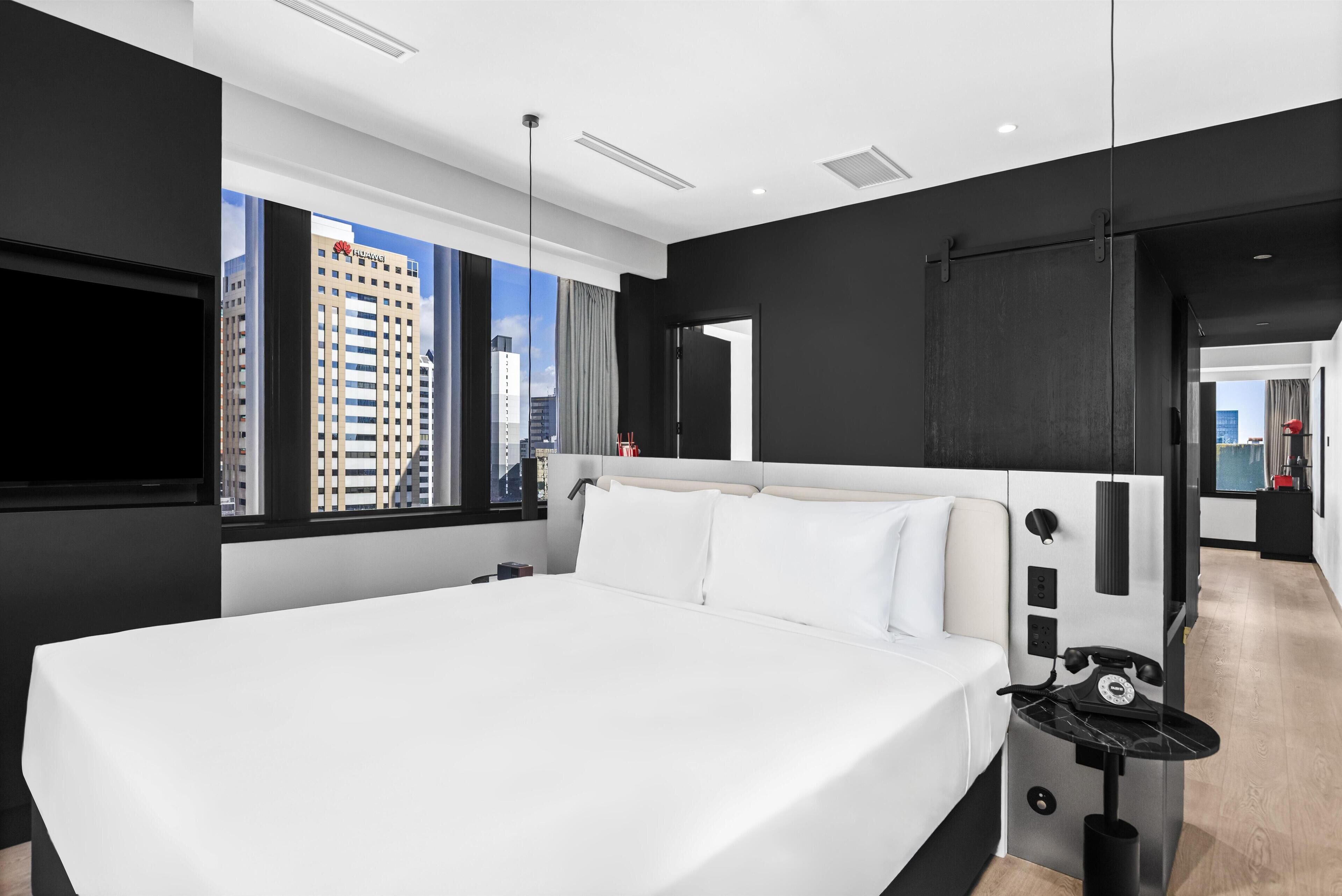 A modern hotel room with a large bed, a TV, a desk with a telephone, and a view of the cityscape.