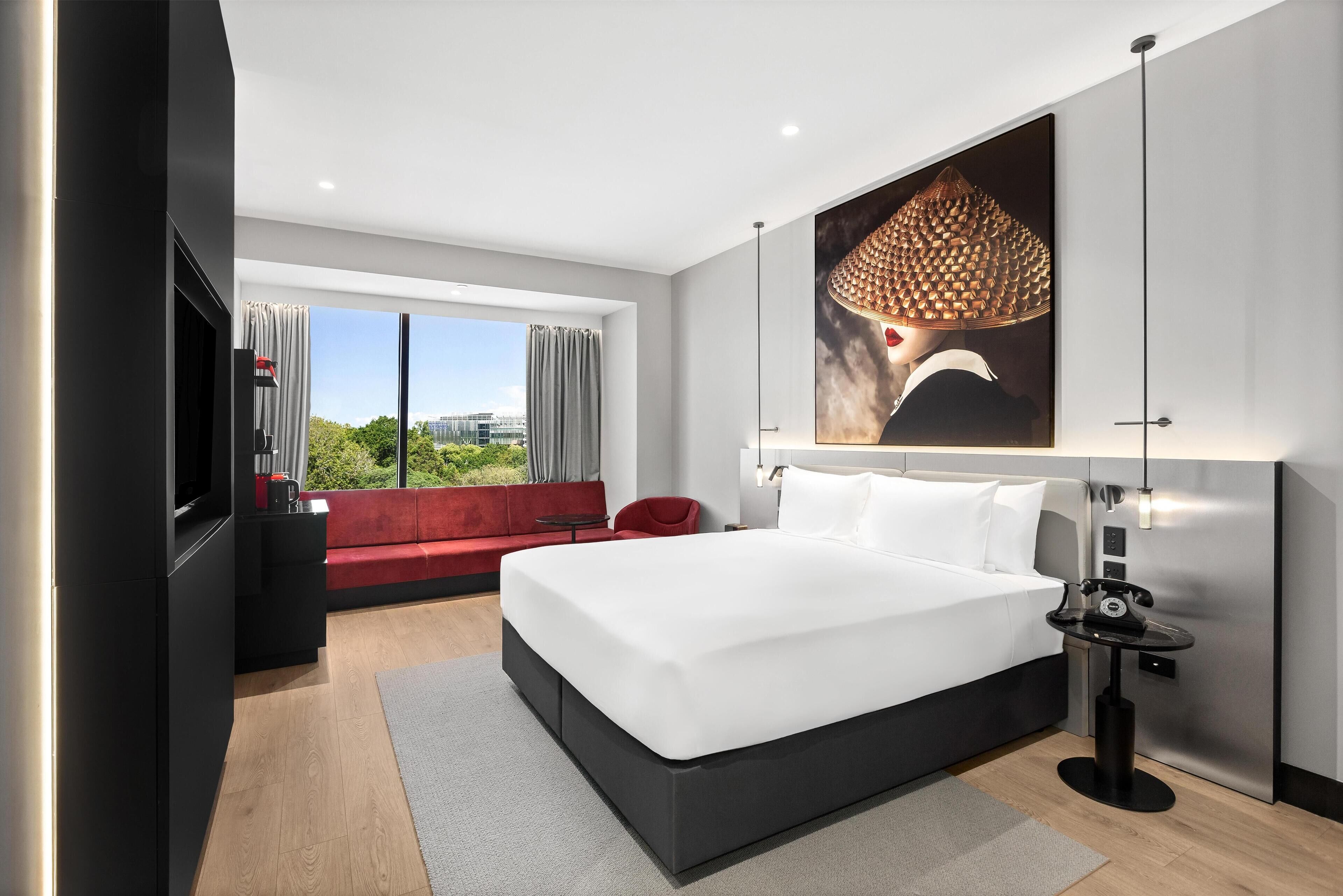 A modern hotel room with a large bed, a red sofa, a small table, and a large window offering a city view.