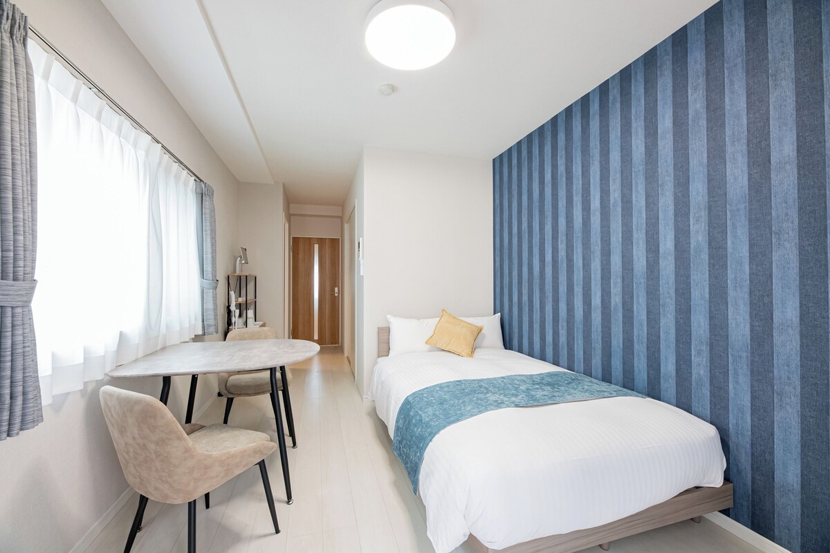 A hotel room with a bed, a small table with chairs, and a view of a hallway.