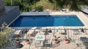 En swimmingpool med solstolar och bord, omgiven av grönska och en klar himmel.