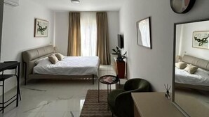 Ett modernt hotellrum med en säng, ett skrivbord, en stol, ett litet bord, en krukväxt och en spegel.