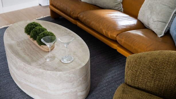 Una mesa de centro redonda de mármol con una pequeña planta en maceta y dos copas de vino, un sofá de cuero marrón y una alfombra oscura en una sala de estar.