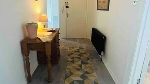 A hallway with a patterned rug, a wooden table with a lamp, a radiator, and a door at the end.
