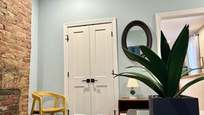 A room with a brick wall, a white door, a wooden chair, a potted plant, a mirror, and a lamp. - Cozy Louisville Gem Mins from U of L Hospital & Downtown (Louisville)