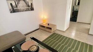 Een moderne woonkamer met een bank, een salontafel met glazen blad en decoratieve objecten, een schilderij aan de muur, een klein houten bijzettafeltje met een lamp, en een groen gestreept vloerkleed.