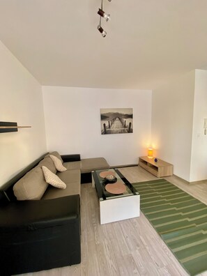 Ein modernes Wohnzimmer mit einer Eckcouch, einem Glastisch, Holzboden, einem grün gestreiften Teppich, einem Wandbild und einem Nachttisch mit Lampe.