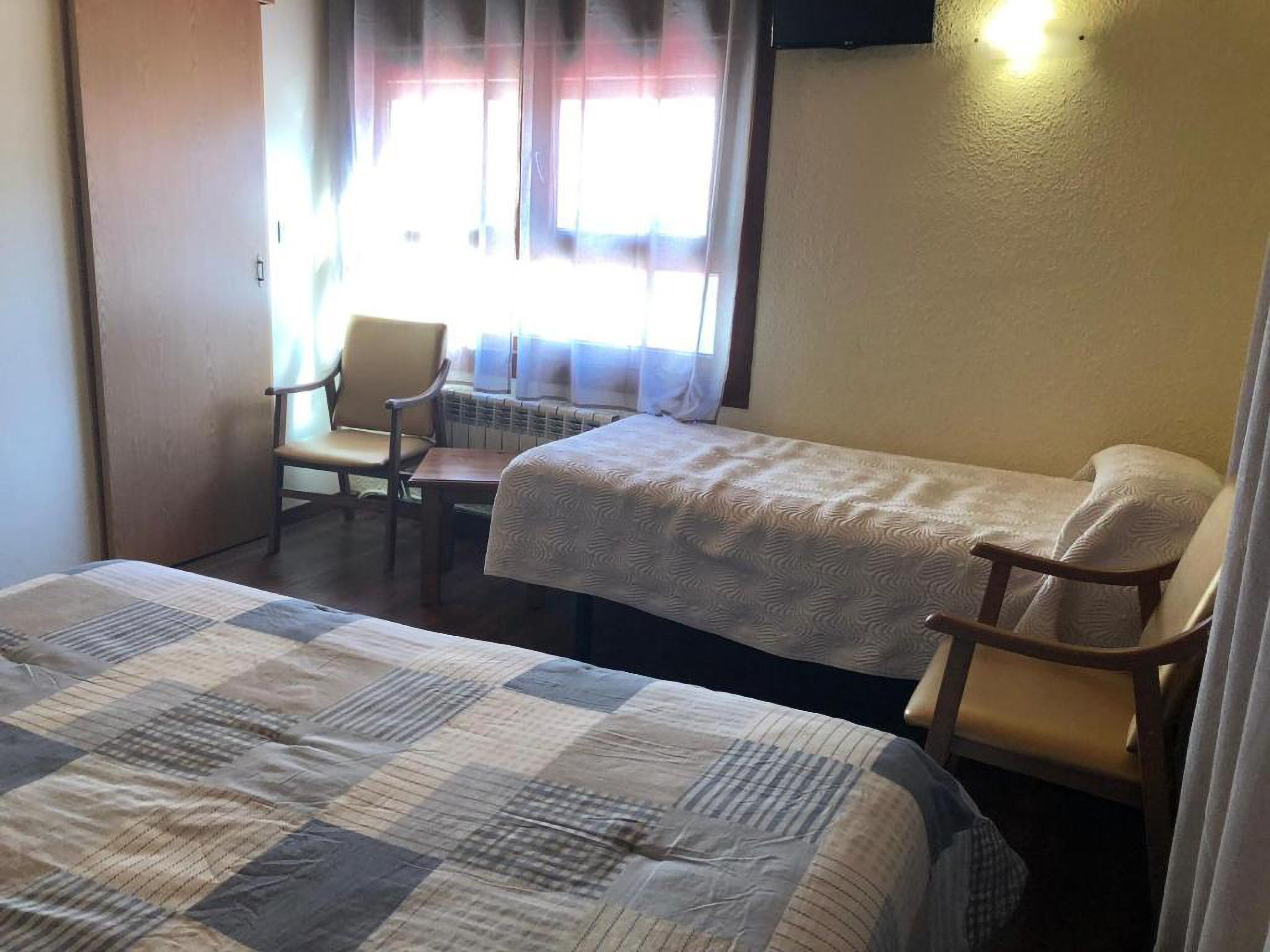 Una habitació senzilla amb dos llits, una cadira i una finestra amb cortines.