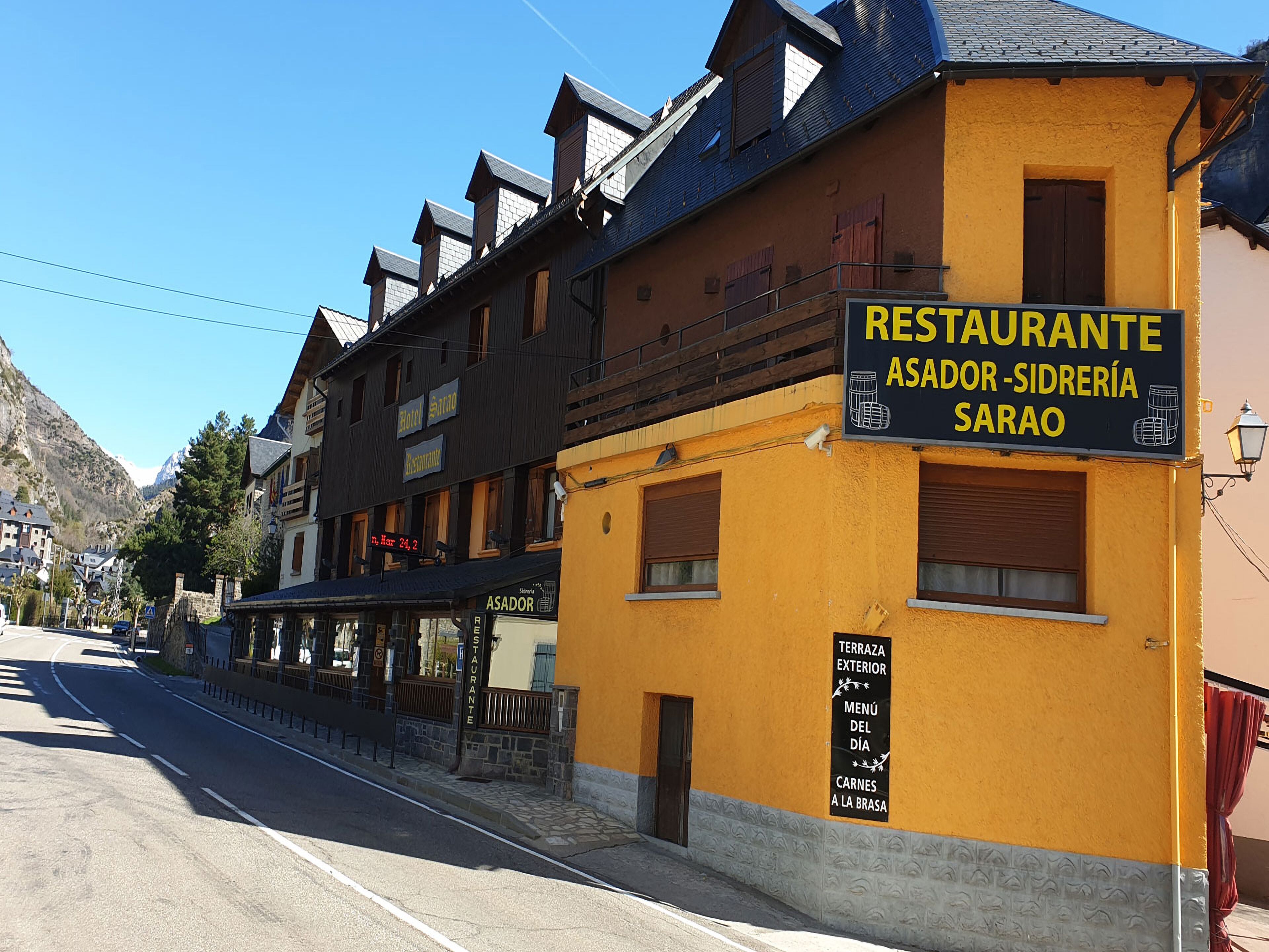 Un bâtiment jaune avec une enseigne indiquant « Restaurante Asador-Sidrería Sarao », doté d’une terrasse extérieure, proposant un menu quotidien et servant de la viande.