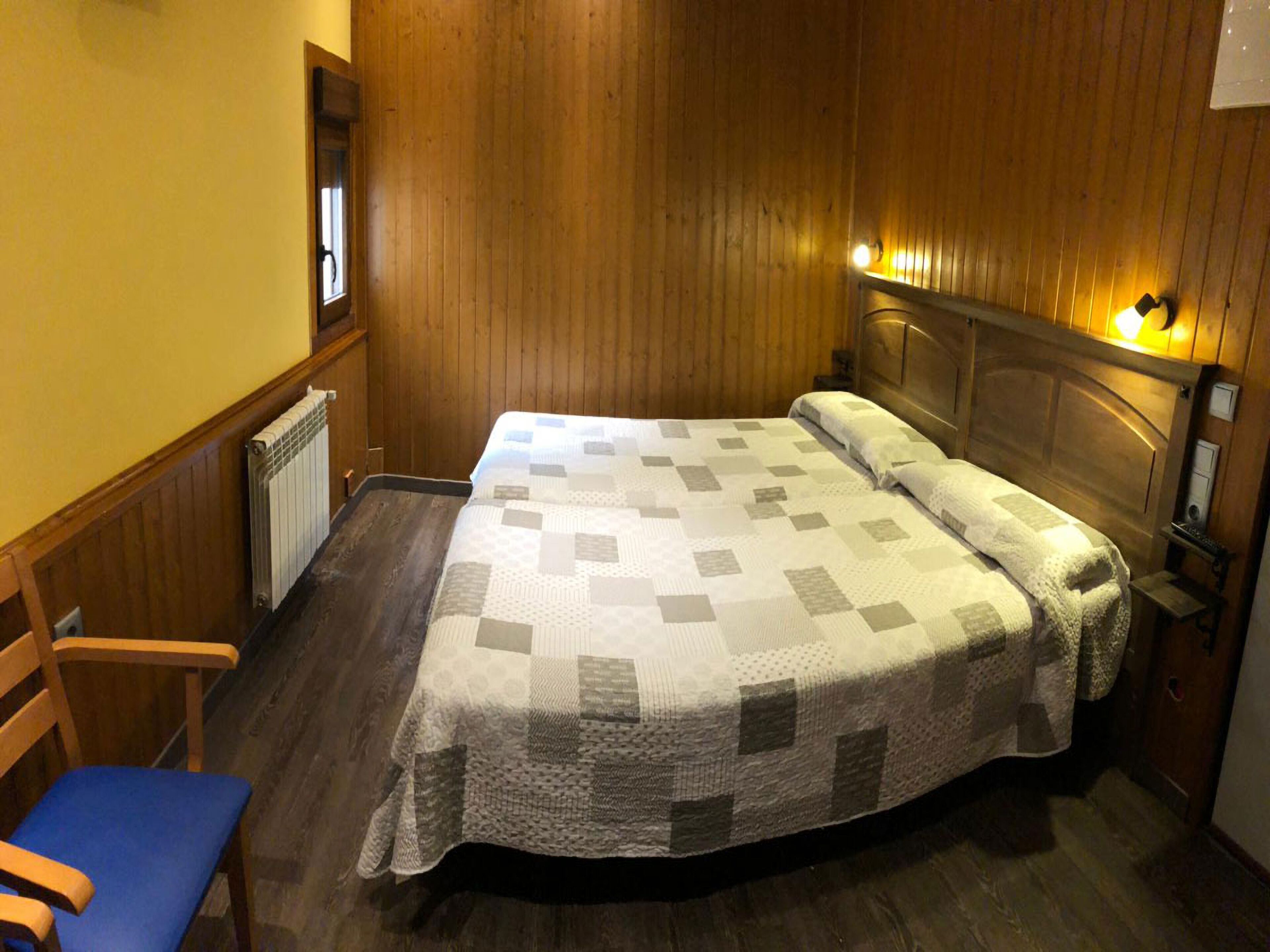 A single bed with a quilt, a wooden chair, a radiator, and a wall-mounted light fixture.