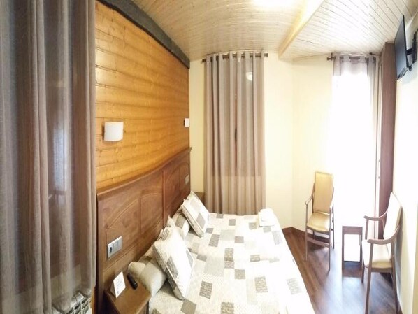 Una habitación de hotel con cabecera de madera, cama con colcha a cuadros, una silla y una ventana con cortinas.