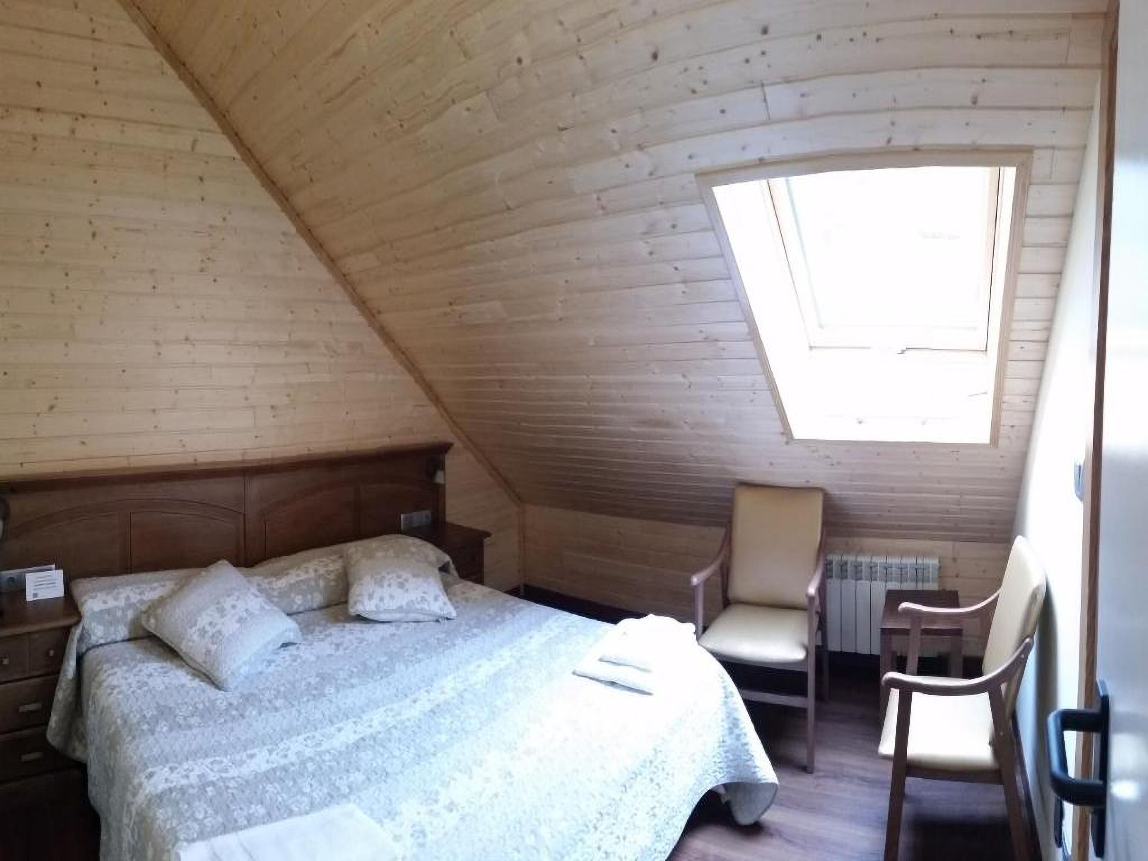 A bedroom with a sloped ceiling, a bed with a patterned bedspread, a chair, and a small window.