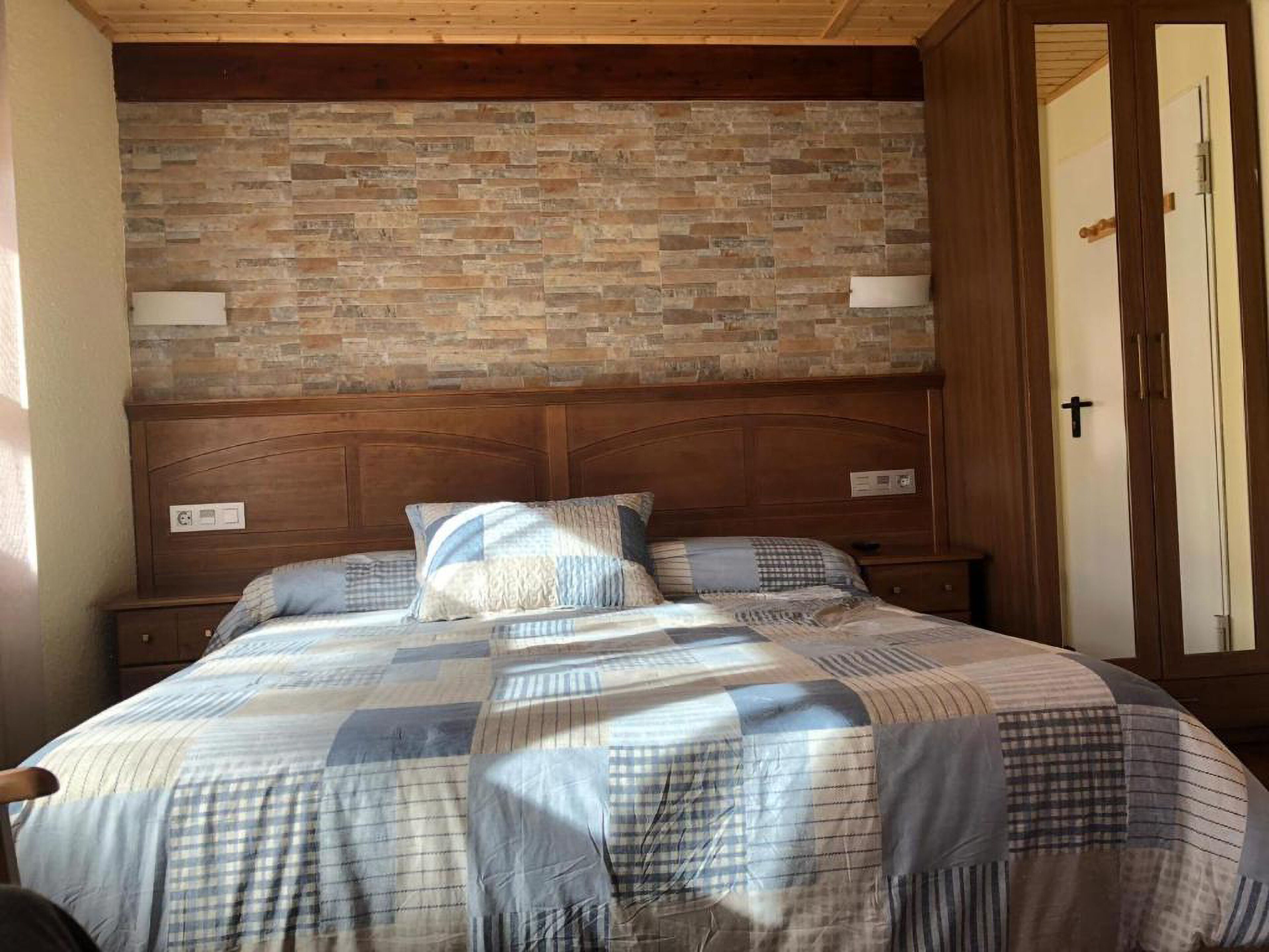A bedroom with a brick-patterned headboard, a bed with a checkered bedspread, and a wooden nightstand with a lamp.
