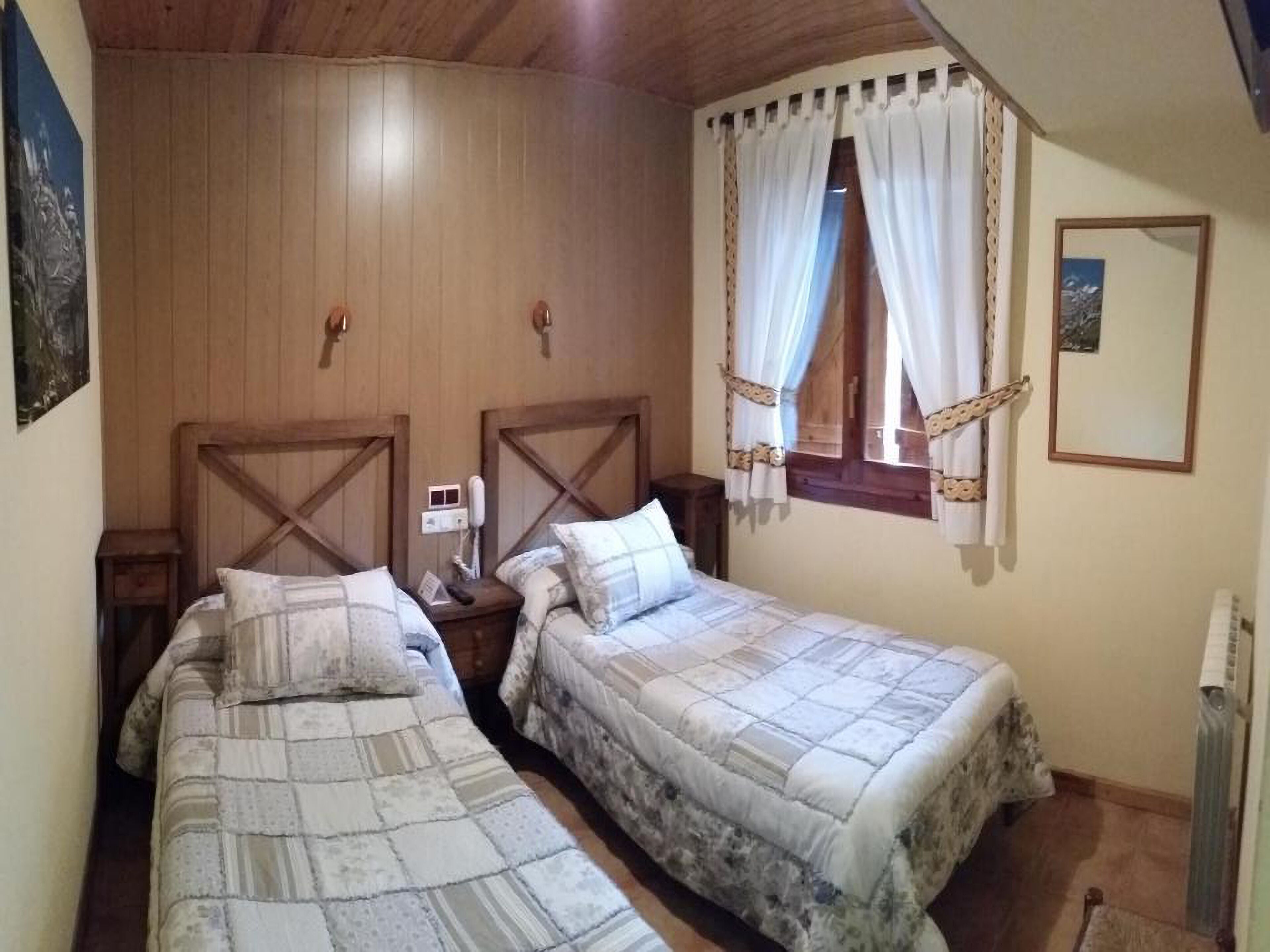 Ein Zimmer mit zwei Einzelbetten, Holz-Kopfteilen, einem kleinen Nachttisch mit Lampe, einem Fenster mit Vorhängen und einem Heizkörper.