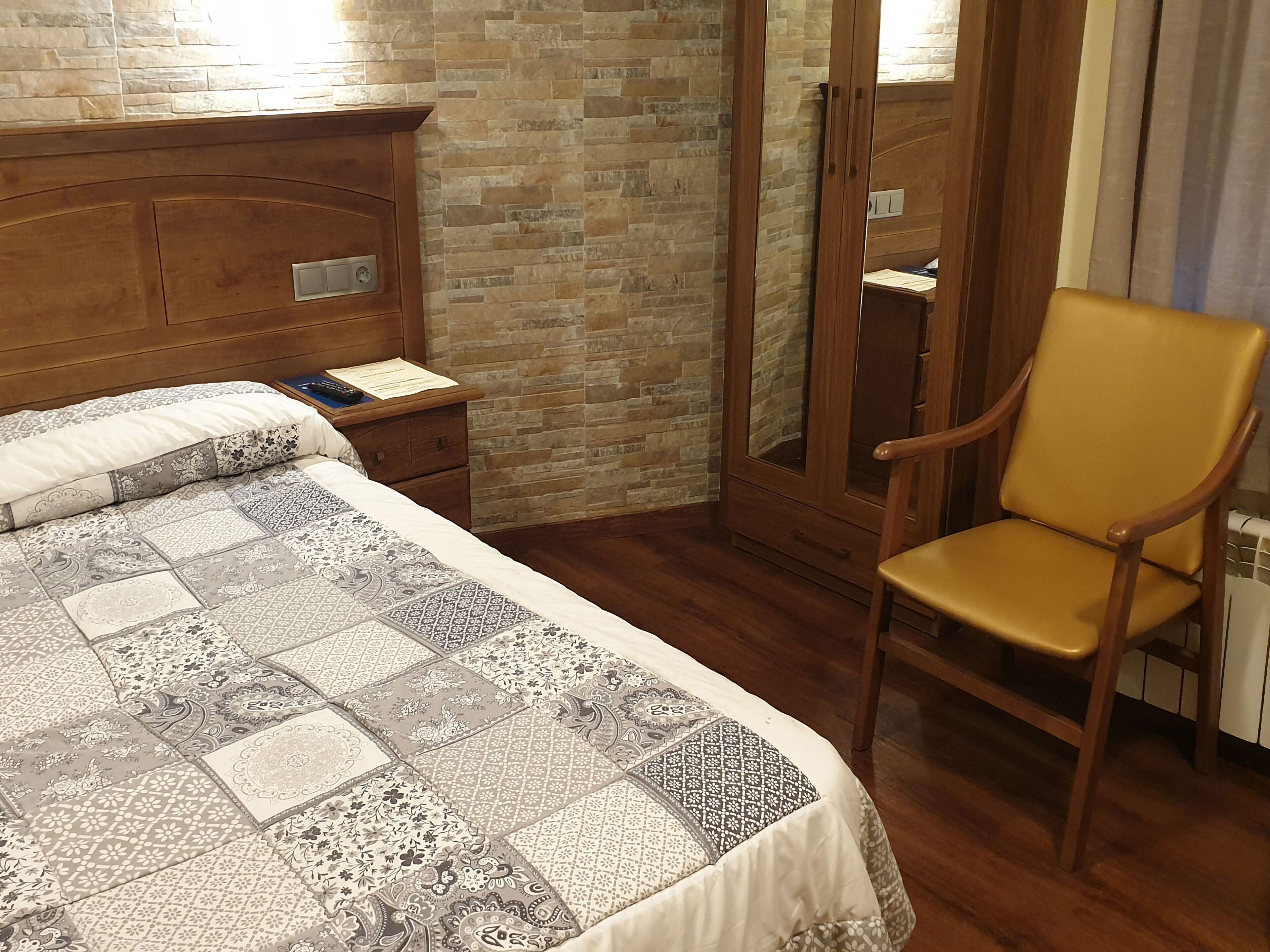 A bedroom with a wooden headboard, a quilted bedspread, a wooden nightstand, a chair, and a stone-textured wall.