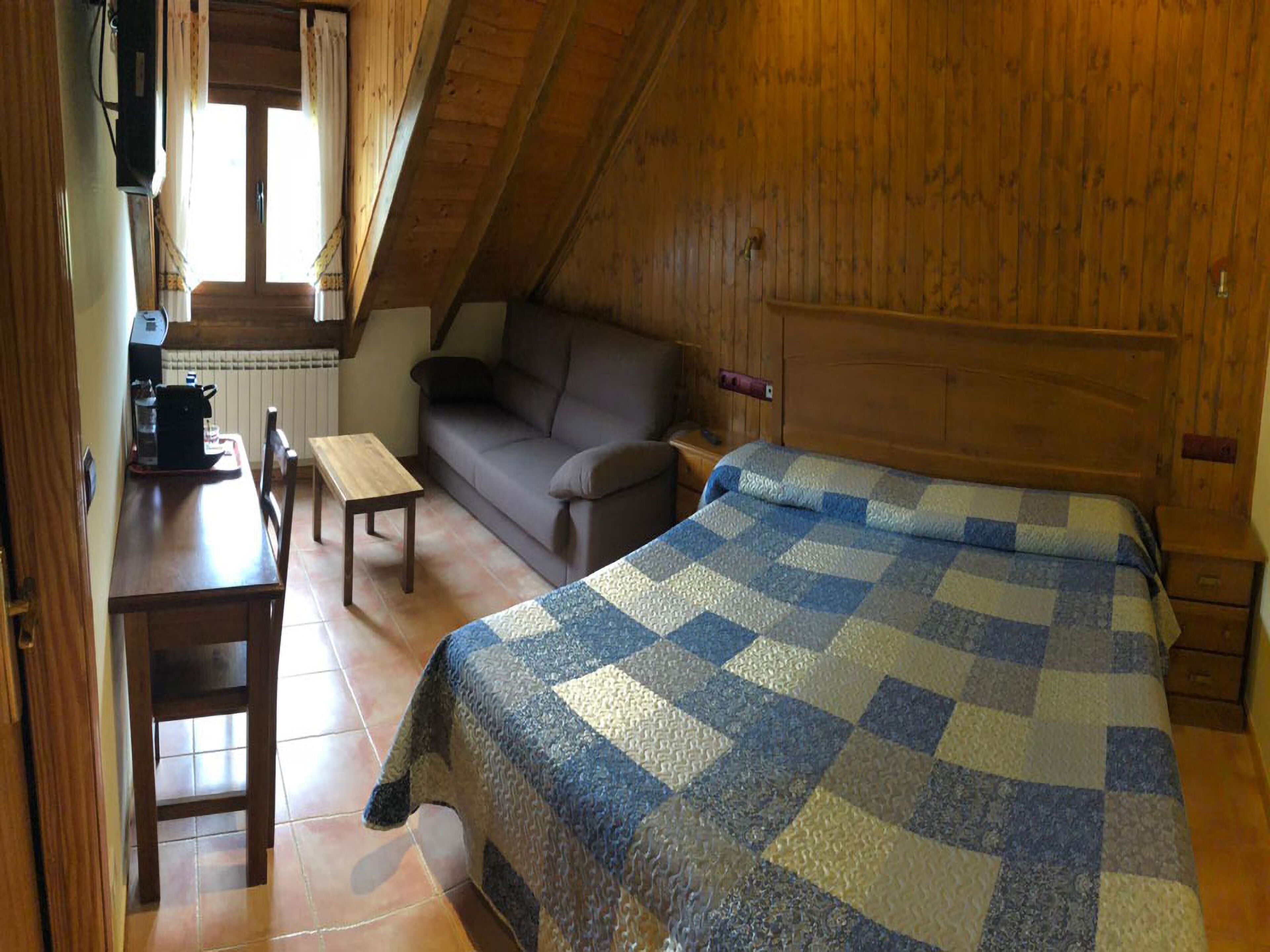 A cozy attic room with a wooden sloped ceiling, a bed with a checkered quilt, a sofa, a small table, and a chair.