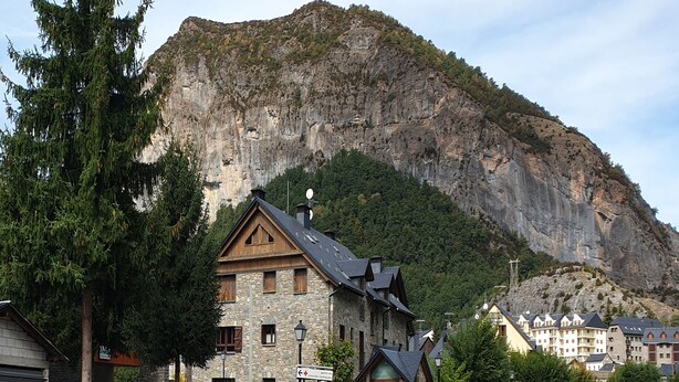 A stone building with a wooden facade, a mountain backdrop, and a street with parked cars. - Hotel Sarao 3000 (Escarrilla)
