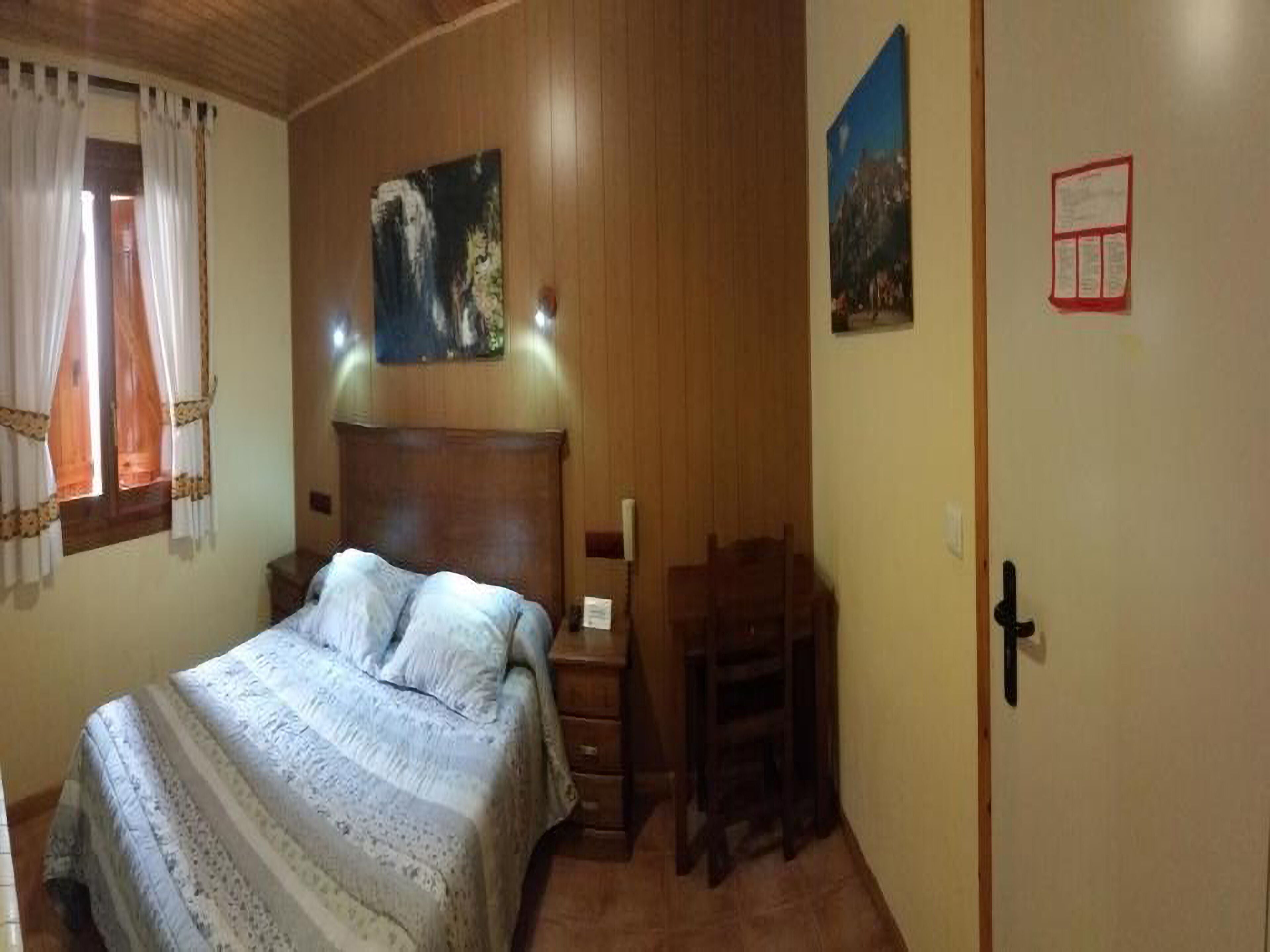 A bedroom with a wooden headboard, a bed with a patterned bedspread, a nightstand, a chair, and a door with a red sign.