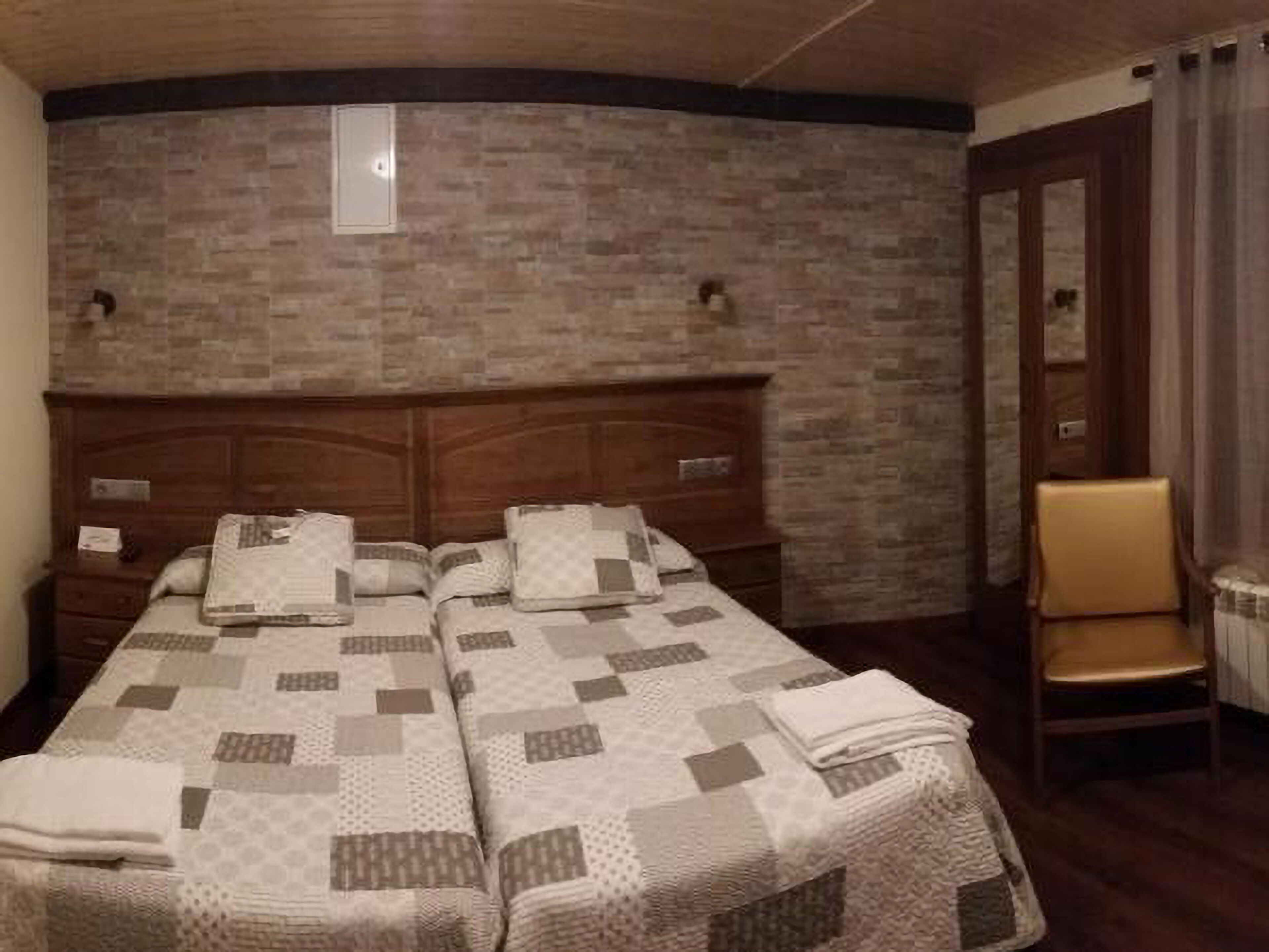 A hotel room with a brick-patterned wall, a wooden headboard, two beds with quilts, a wooden nightstand, a chair, and a door with curtains.