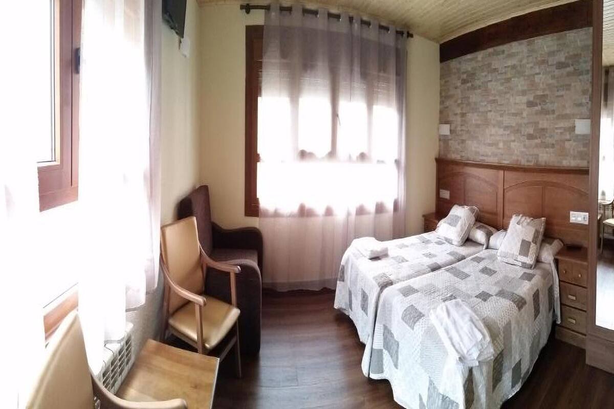 A bedroom with two beds, a wooden chair, a small table, and a window with curtains.