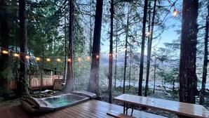 Une terrasse en bois avec un jacuzzi et une table de pique-nique, entourée de grands arbres et illuminée de guirlandes lumineuses.