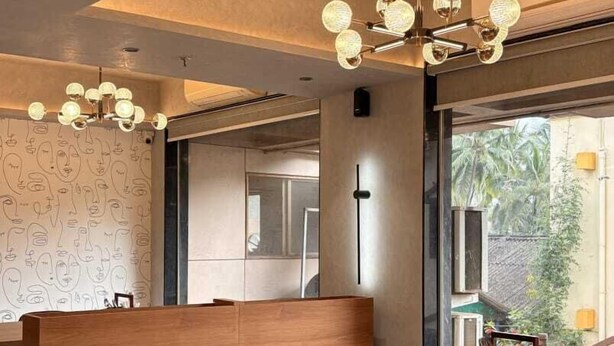 A modern dining area with a wooden table and chairs, a unique ceiling light fixture, and a view of the outdoors. - Avron Beach Resort (Daman)
