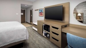 A hotel room with a bed, a TV mounted on a wooden cabinet, a desk with drawers, a lamp, and a painting on the wall. - Near Logan Airport + Free Breakfast. Shuttle. Gym. (Chelsea)