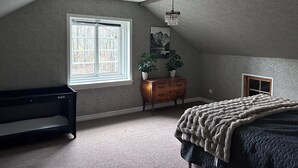 Une chambre avec un plafond en pente, une grande fenêtre, une commode, un lit et un luminaire suspendu.