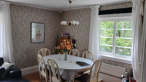 Un coin repas comprenant une table ronde, six chaises, un lustre, une fenêtre donnant sur un espace verdoyant et un radiateur.
