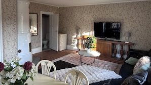 Un salon avec un téléviseur, une table à manger avec des chaises, un canapé, une petite table avec une plante, et une salle de bain visible à travers une porte ouverte.