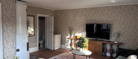 A living room with a TV, a dining table with chairs, a sofa, a small table with a plant, and a bathroom visible through an open door.