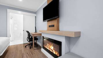 A modern hotel room with a flat-screen TV mounted above a fireplace, a wooden desk, and a black office chair.