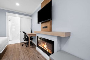 Een moderne hotelkamer met een flatscreen televisie boven een open haard, een houten bureau en een zwarte bureaustoel.